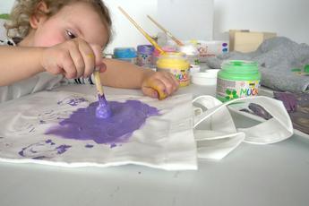 Fragen Antworten Tipps FAQ MUCKI Stoff-Fingerfarbe für Kinder ab 2 Jahren Textilfarbe Kind stempelt mit lila MUCKI Stoff-Fingerfarbe auf einer weißen Baumwolltasche